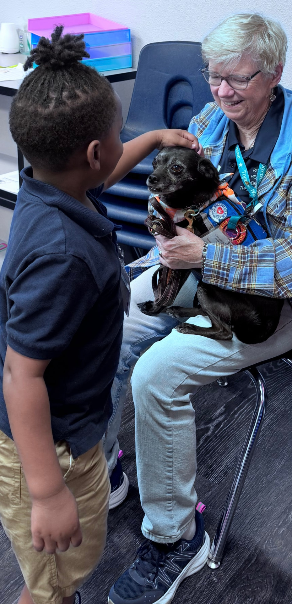 A young student reaching to pet Luna, a small black Chihuahua mix therapy dog, held by her handler during a school visit