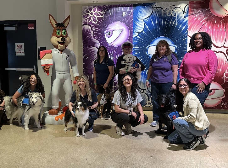 Paws 4 Hearts volunteers with their therapy dogs posing in front of a colorful mural and team mascot