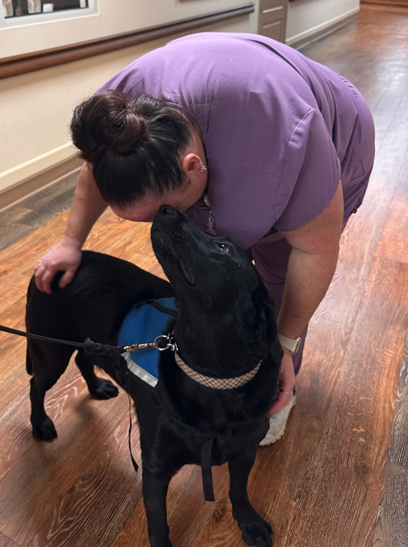 A staff member in purple scrubs leaning down to hug Duke, a black Labrador therapy dog in a blue vest, indoors on a wood floor