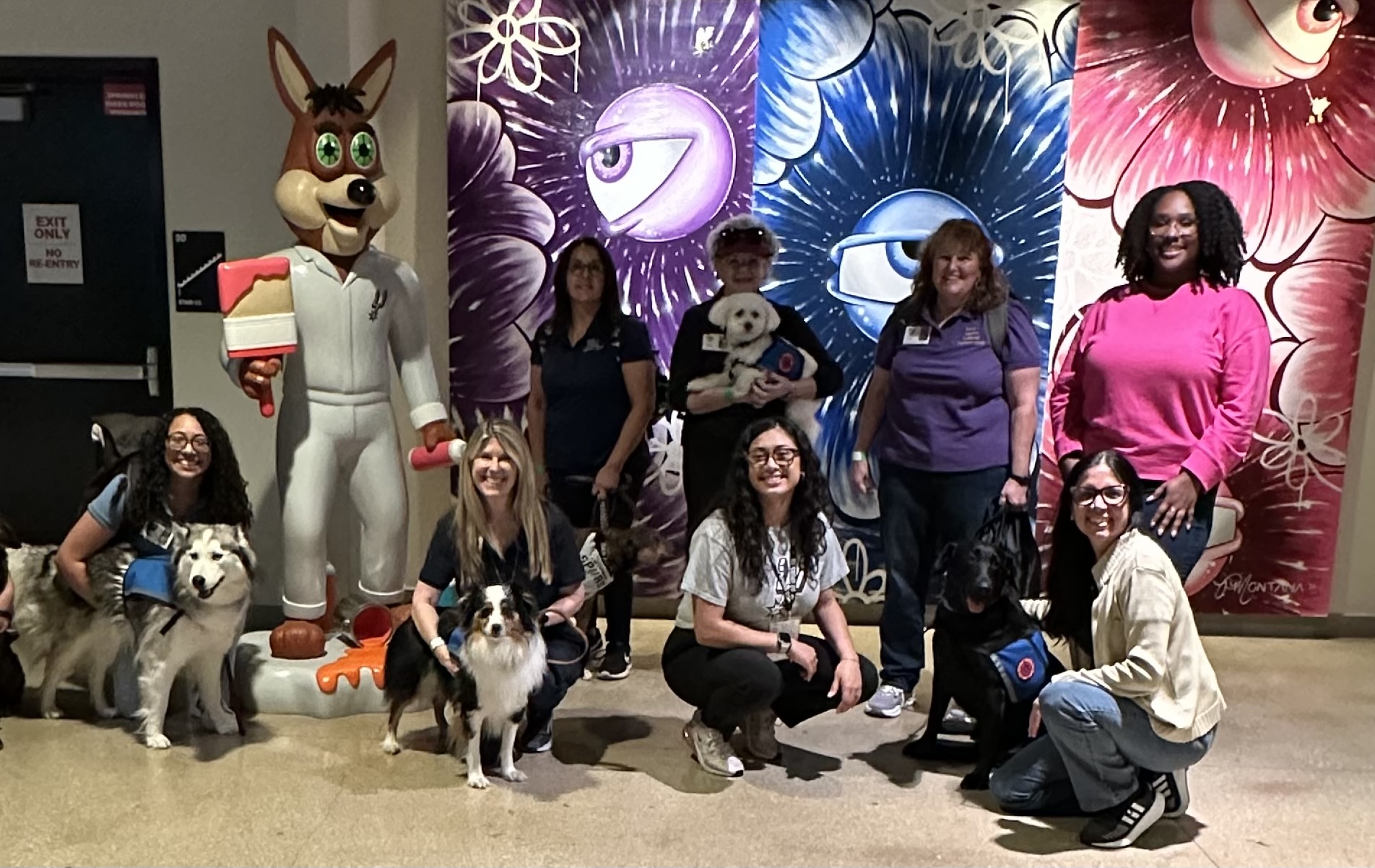 Volunteers with therapy dogs posing with the Spurs Coyote mascot at an indoor community event